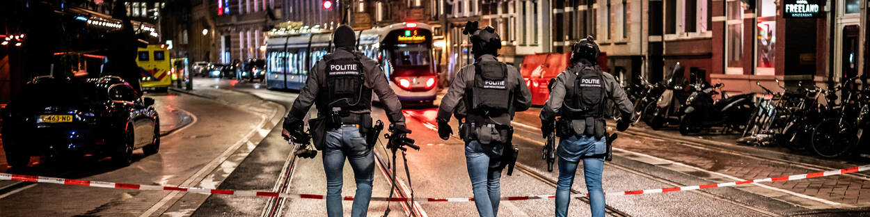 3 agenten arrestatieteam op hun rug gezien lopen in een winkelstraat die met lint is afgezet, in de avond, tram rijdt voorbij