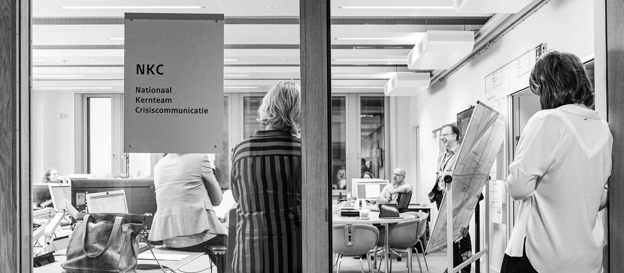 Inkijkje door deur met personen op de rug gezien in een kantoorruimte met op het raam een bord Nationaal Kernteam Crisiscommunicatie