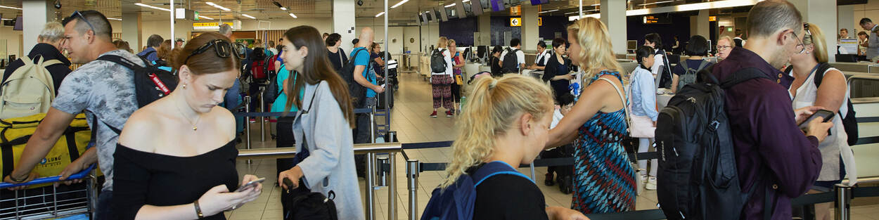 Diverse mensen lopen door elkaar met karretjes, tassen, kijkend op hun telefoon of staan in de balie bij Schiphol