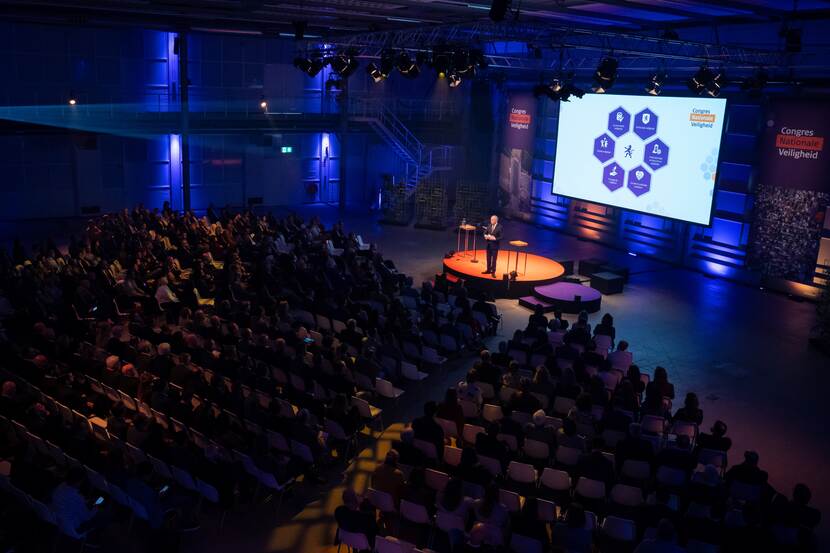 Grote zaal vol mensen, donker, met op podium een spreker en op de achtergrond een presentatie met een grafiek in de spotlight