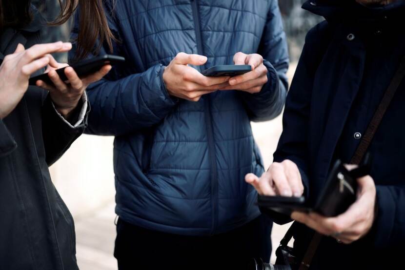 3 jongeren met winterjassen aan waarvan alleen hun handen te zien zijn waar ze hun mobiel in vasthouden en op tikken, swipen