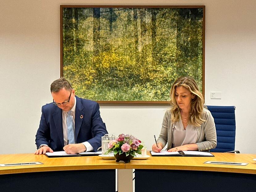 Dilan Yesilguz zit naast een man aan lange tafel, ze ondertekenen beiden een document op de achtergrond is mooie foto van een bos te zien
