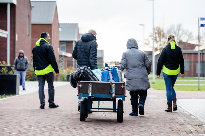 Man en vrouw lopen in een straatje in winterjas en met karretje met spullen worden begeleid door 2 personen van een AZC, allen van achter gefotografeerd