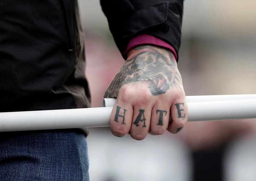Een man houdt een stok vast. Op zijn vingers staat het woord HATE getatoeëerd.