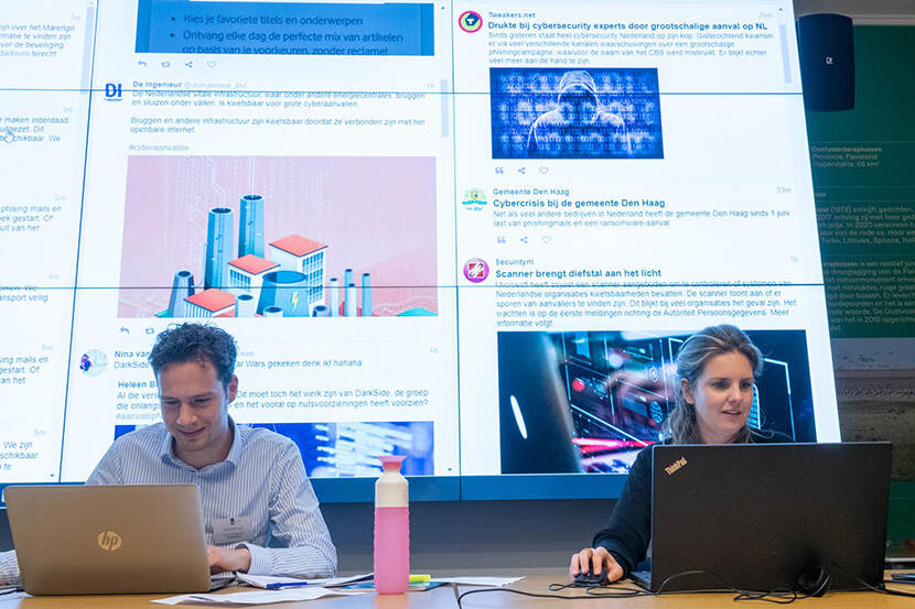 2 personen achter een laptop met op achtergrond groot scherm met een website erop
