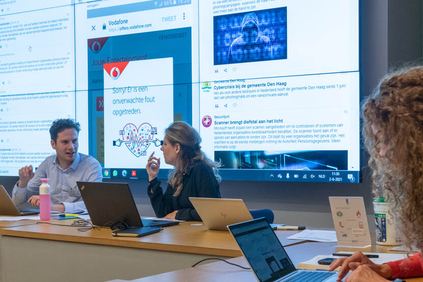 2 personen achter een laptop met erachter een groot scherm met een website erop