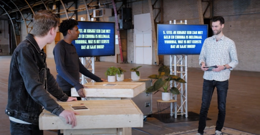 Youtube stilstaand beeld uit gastles studio corona met 2 personen achter een desk en een leraar ervoor met op beeldscherm tekst van de les